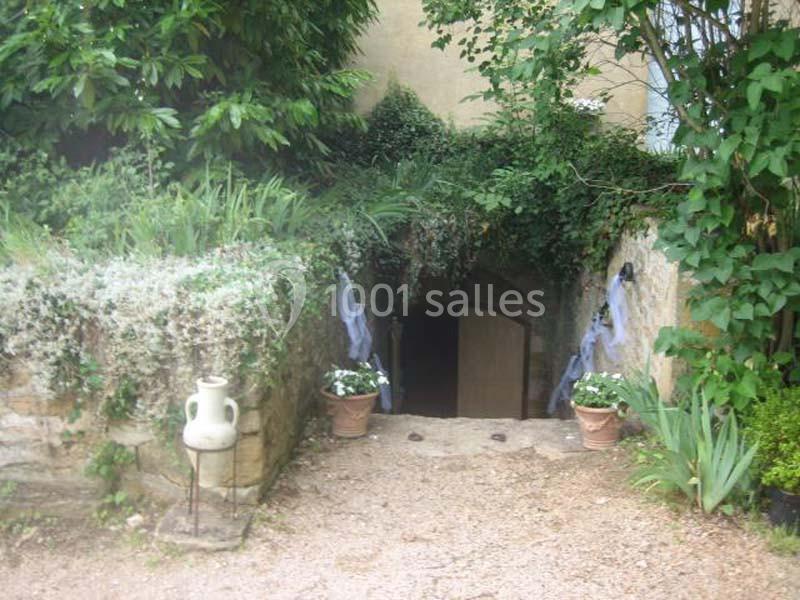 Entrée voûtée d'une cave entourée de végétation, avec des pots de fleurs et un chemin en gravier.