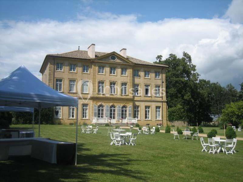 Façade d'un grand bâtiment ancien entouré d'un jardin avec tables et chaises blanches disposées sur la pelouse.
