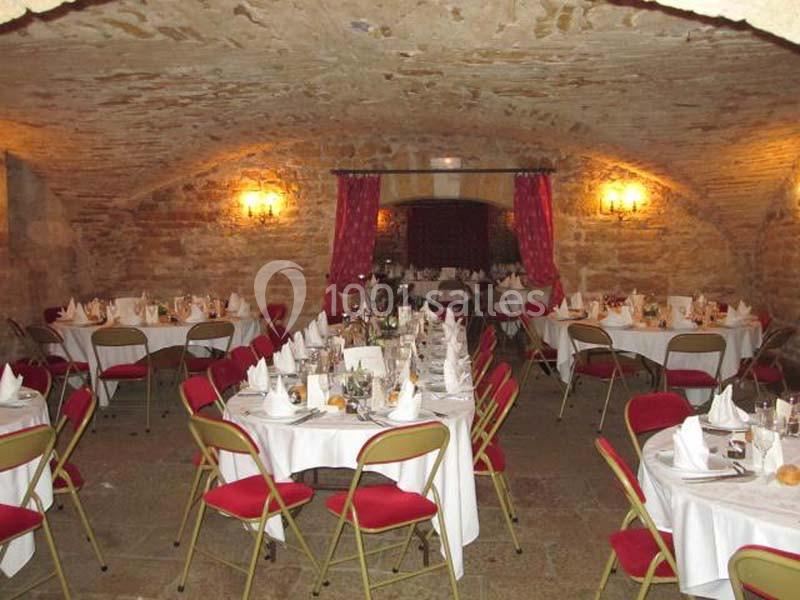 Salle voûtée en pierre avec tables dressées pour un repas, chaises rouges et éclairage chaleureux.