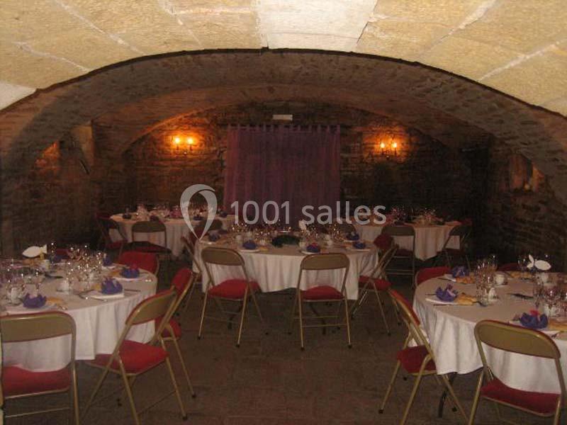 Salle voûtée en pierre aménagée pour un repas, avec tables rondes dressées et chaises pliantes rouges.