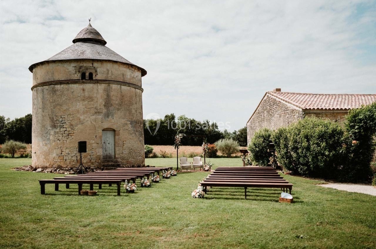 Un jardin avec un pigeonnier ancien, des bancs en bois alignés et des décorations florales pour une cérémonie en plein air.