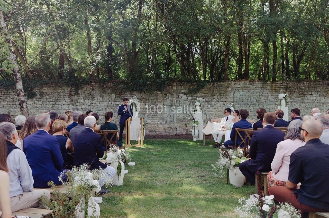 Cérémonie de mariage en plein air avec des invités assis, un officiant et les mariés devant un mur en pierre.