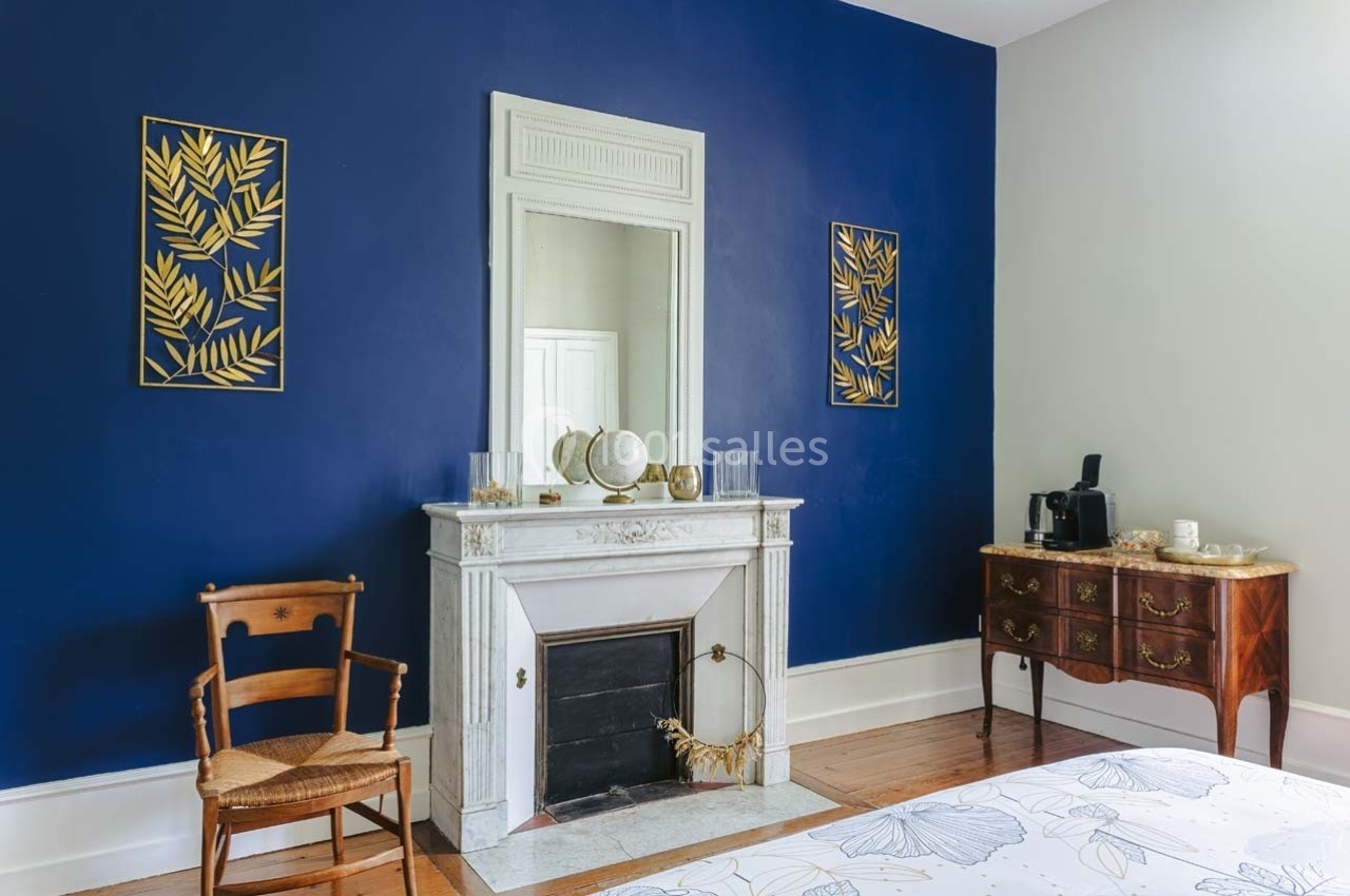 Cheminée blanche avec miroir, mur bleu décoré de cadres dorés, meuble en bois et chaise dans une chambre lumineuse.