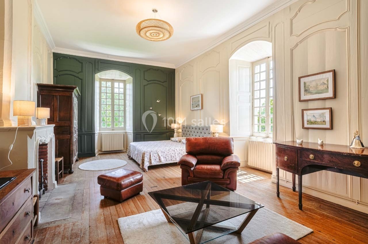 Chambre lumineuse avec lit double, fauteuils en cuir, meubles en bois et grandes fenêtres laissant entrer la lumière…