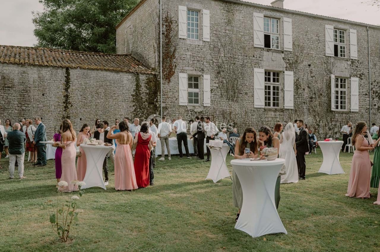 Groupe de personnes rassemblées dans un jardin devant un bâtiment en pierre, autour de tables hautes.