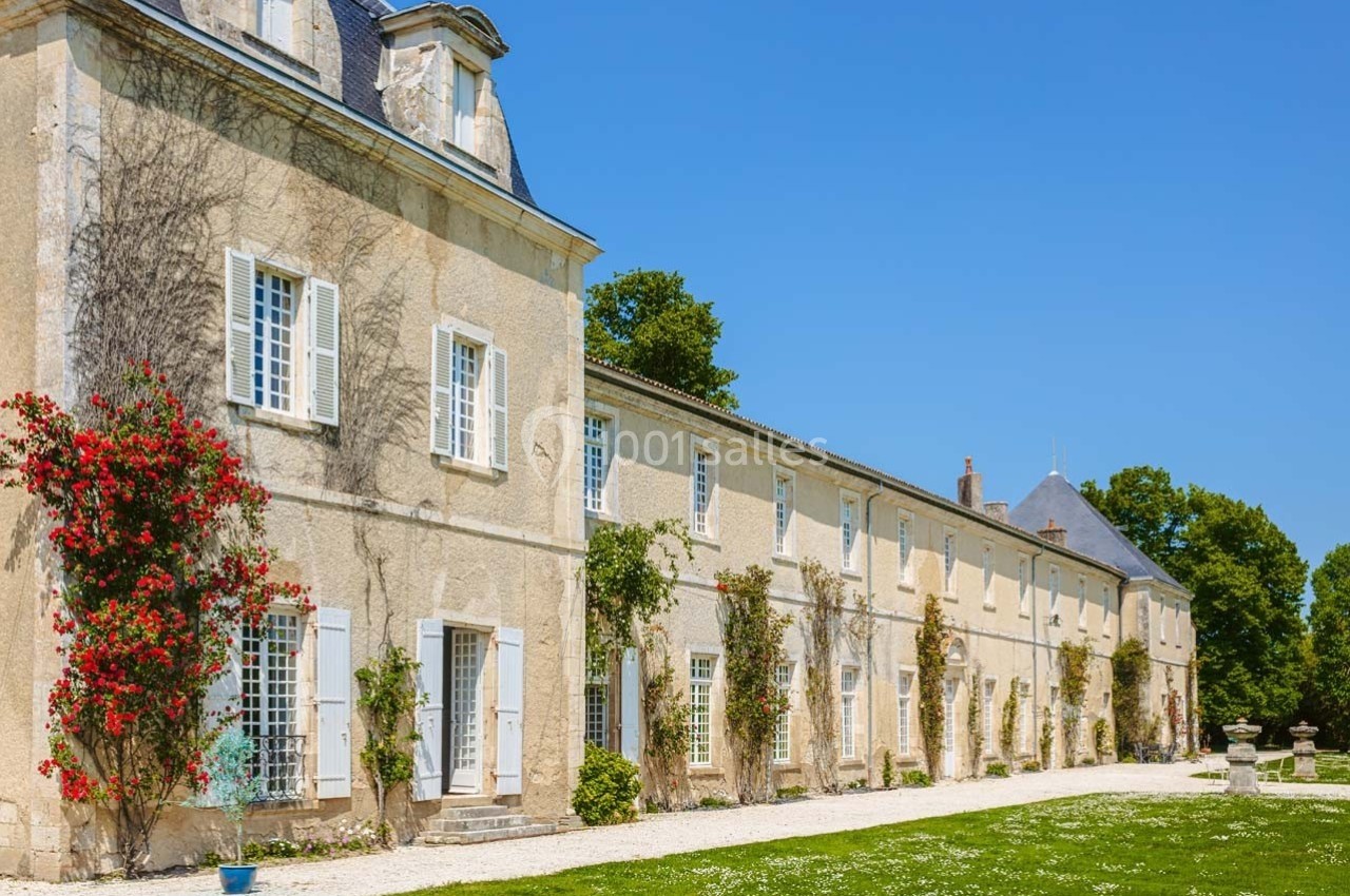 Façade d'un grand bâtiment en pierre avec volets blancs, entouré de végétation et d'un jardin sous un ciel bleu.