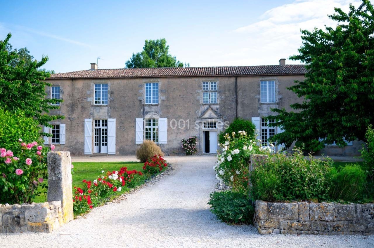 Façade d'une maison en pierre avec volets blancs, entourée d'un jardin fleuri et d'une allée gravillonnée.