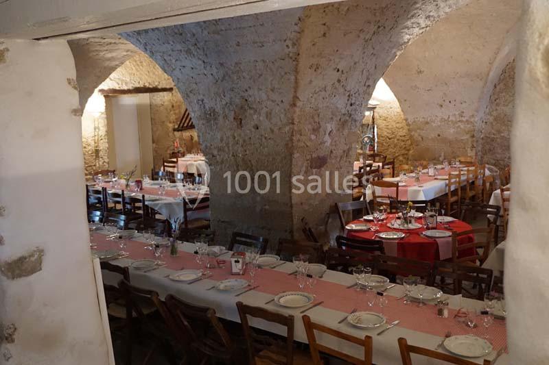 Salle voûtée en pierre aménagée avec de longues tables dressées pour un repas, décorée de nappes et vaisselle.