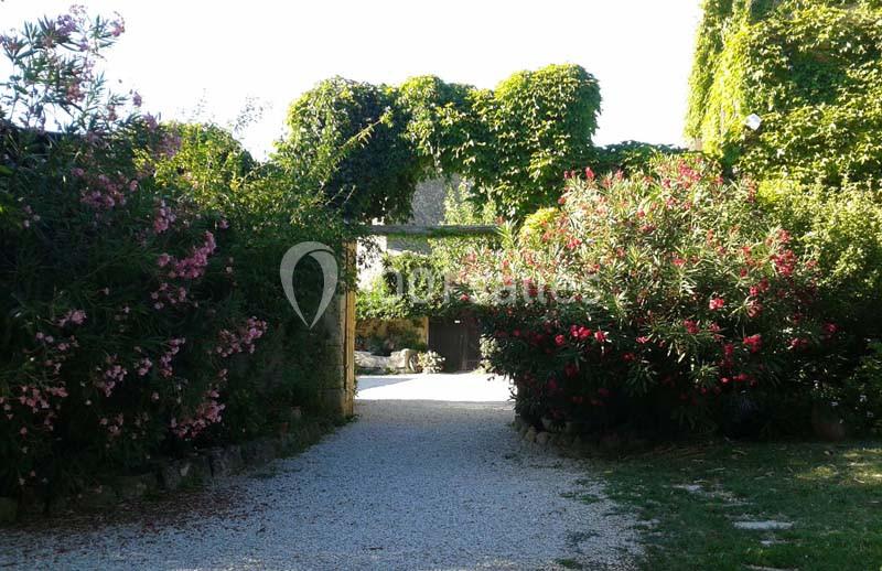Allée gravillonnée bordée de lauriers roses et de végétation luxuriante, menant à un portail en pierre.