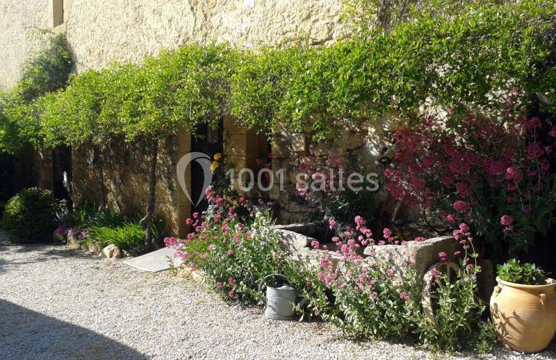 Façade en pierre avec plantes grimpantes, fleurs colorées et arrosoir posé près d'un parterre fleuri.