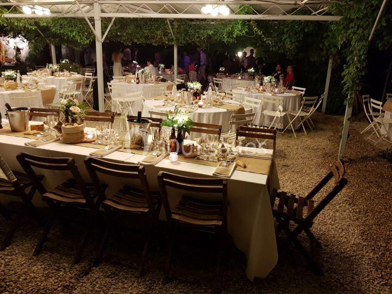 Tables décorées pour un dîner en extérieur, sous une tonnelle éclairée, avec chaises en bois et ambiance conviviale.