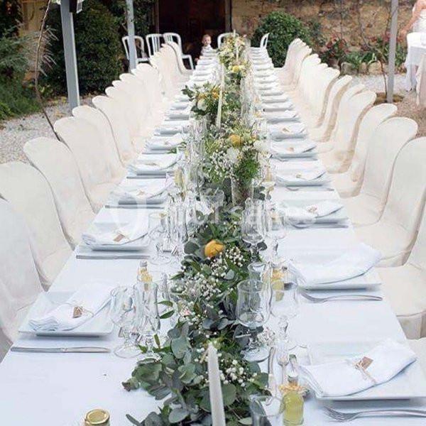 Grande table extérieure décorée de nappes blanches, bougies, fleurs et feuillages, entourée de chaises blanches.