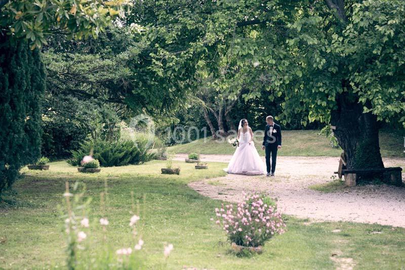 Un couple en tenue de mariage marche dans un jardin verdoyant, entouré d'arbres et de fleurs.