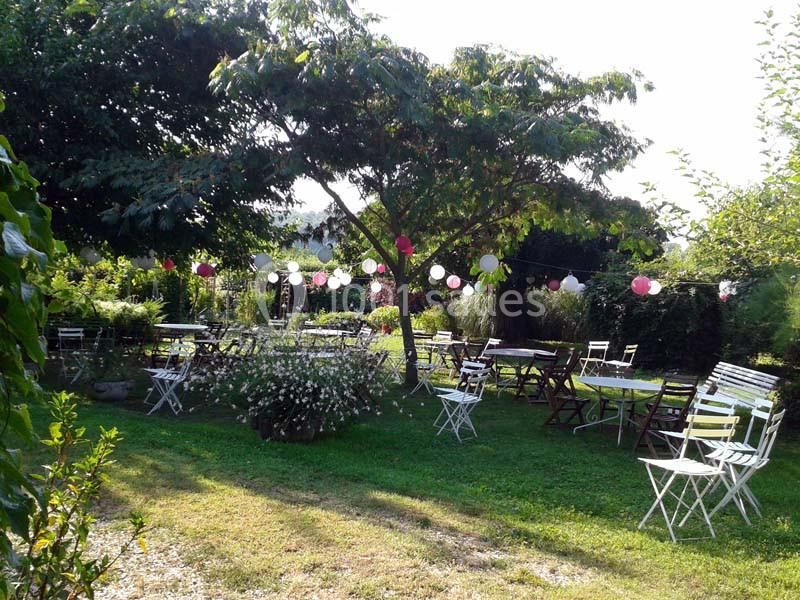 Chaises et tables blanches et en bois disposées dans un jardin verdoyant, décoré de guirlandes lumineuses colorées.