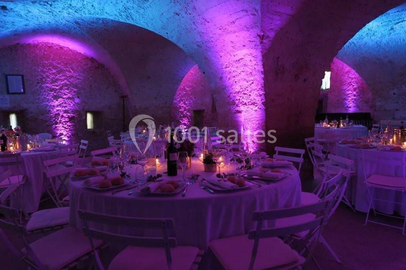 Salle voûtée éclairée de lumières violettes, avec tables rondes décorées pour un dîner ou un événement festif.