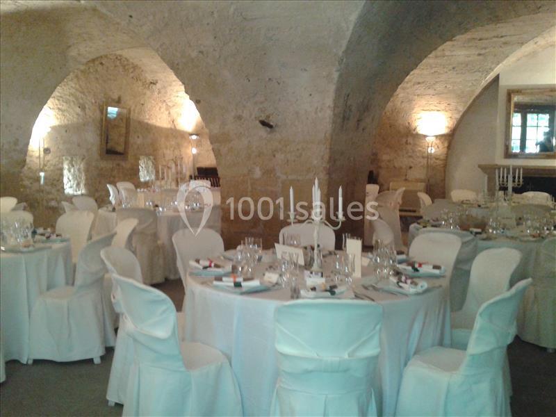 Salle voûtée en pierre aménagée pour un événement, avec tables rondes dressées et chaises couvertes de housses blanches.