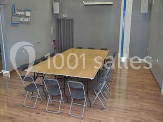 Salle vide avec des murs gris, une table rectangulaire en bois entourée de chaises pliantes sur un sol en parquet.