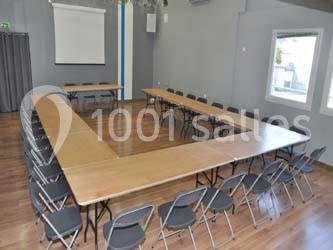 Salle de réunion avec des tables disposées en U et des chaises pliantes, éclairée par des fenêtres et un projecteur visible.