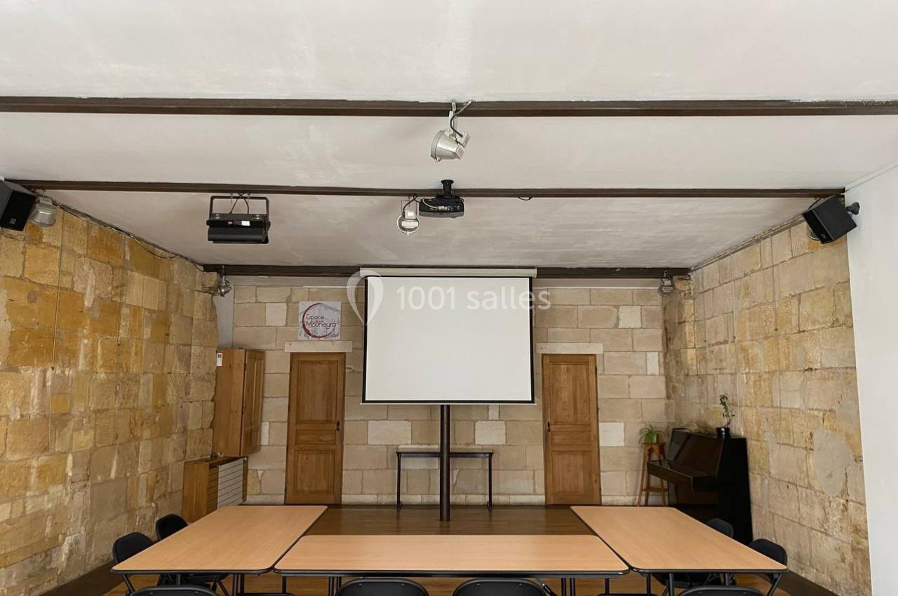 Salle avec murs en pierre, écran de projection, tables disposées en U et chaises noires pliantes.