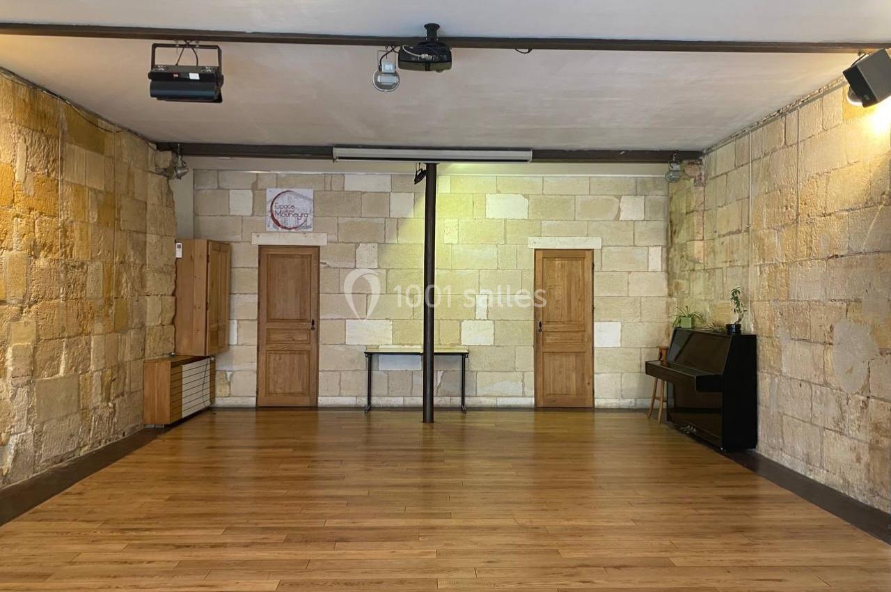 Salle vide avec parquet en bois, murs en pierre, trois portes en bois, un piano noir et des projecteurs au plafond.