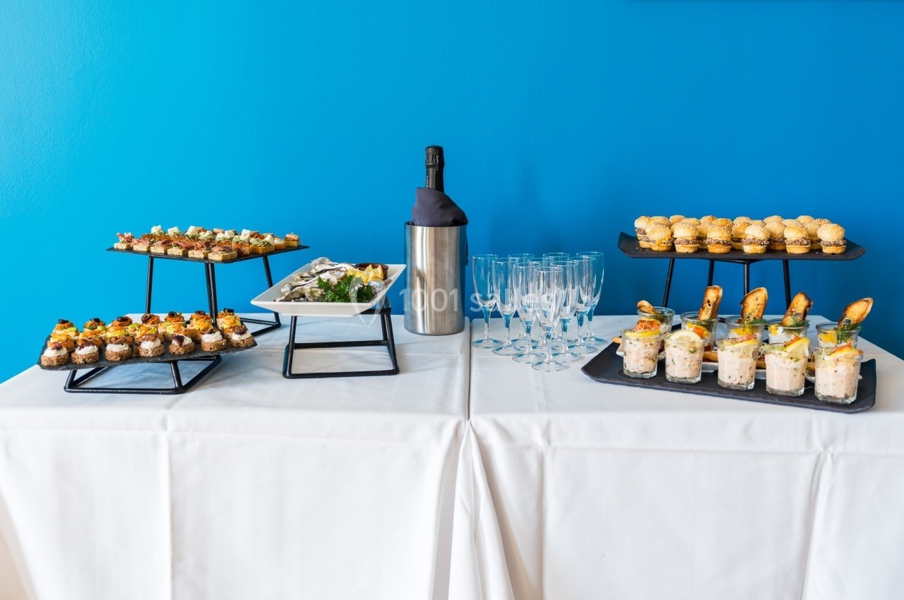 Table dressée avec des amuse-bouches variés, une bouteille de champagne et des flûtes devant un mur bleu.