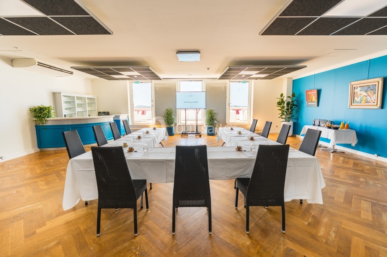 Salle de réunion lumineuse avec des tables disposées en U, des chaises noires et un écran de présentation au centre.