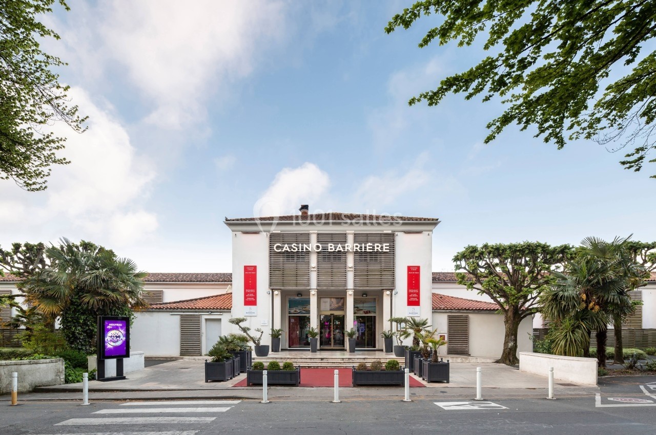 Façade d'un casino avec une entrée principale entourée de végétation et un ciel partiellement nuageux en arrière-plan.
