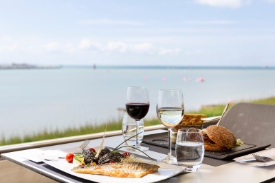 Repas avec poisson, burger, vin et eau servis sur une table en terrasse avec vue sur la mer.