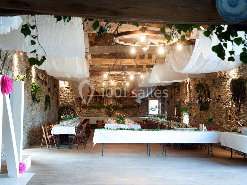 Salle de réception rustique décorée avec des guirlandes, des tables longues et des nappes blanches sous un plafond en bois.