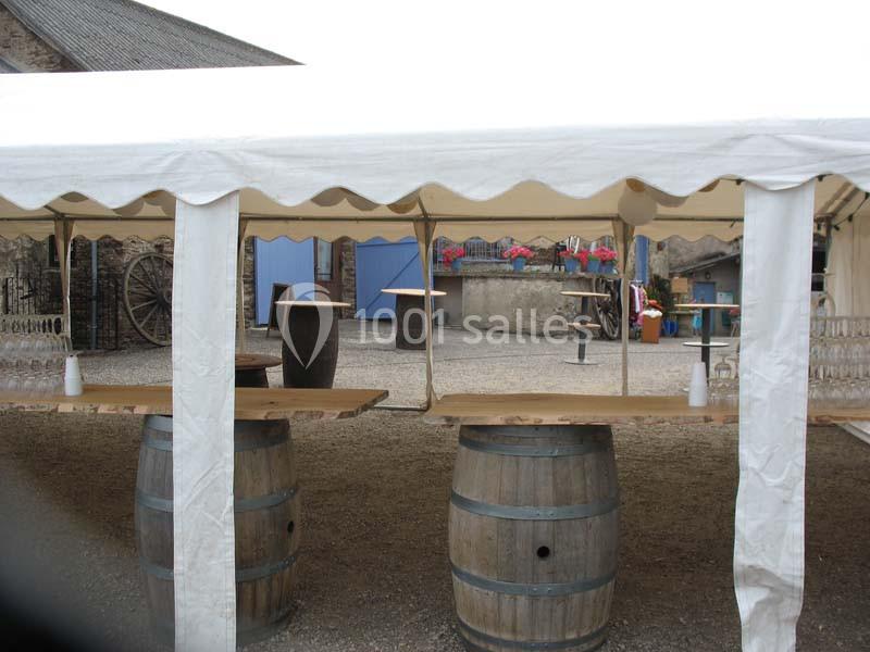 Tonnelles blanches abritant des tables en bois posées sur des tonneaux, dans une cour extérieure avec des fleurs en arrière…