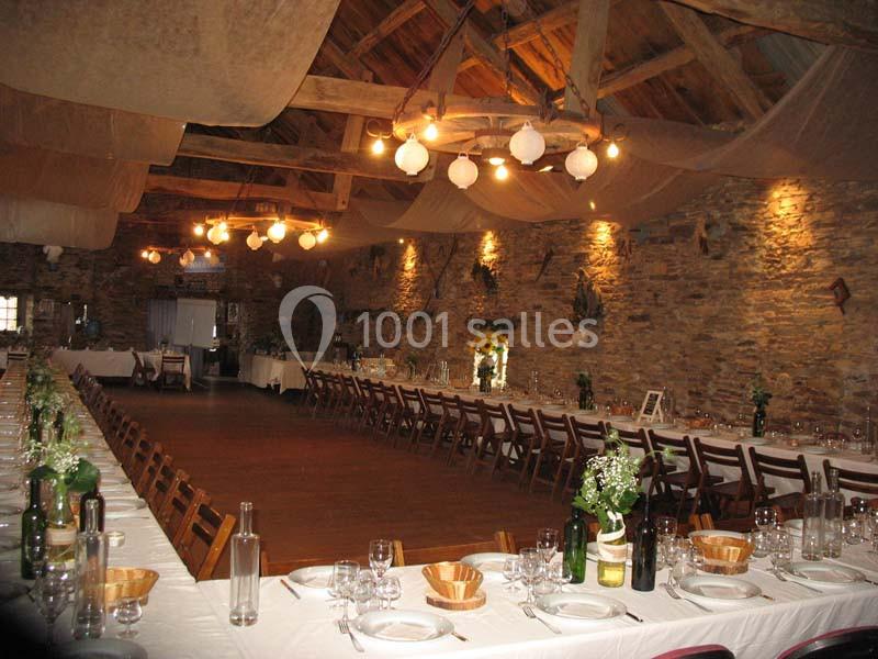 Grande salle rustique avec murs en pierre, tables longues dressées pour un repas, éclairée par des suspensions lumineuses.