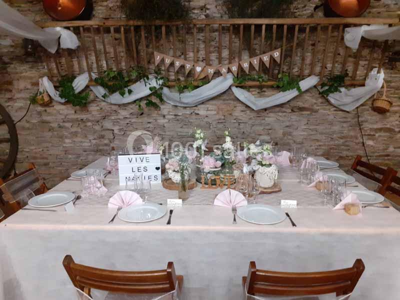 Table décorée pour un mariage avec nappes blanches, vaisselle, fleurs et guirlandes dans une salle rustique.