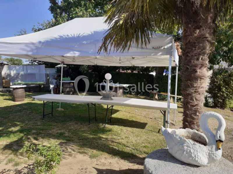 Tente blanche montée dans un jardin avec tables pliantes, une statue de cygne et un palmier au premier plan.