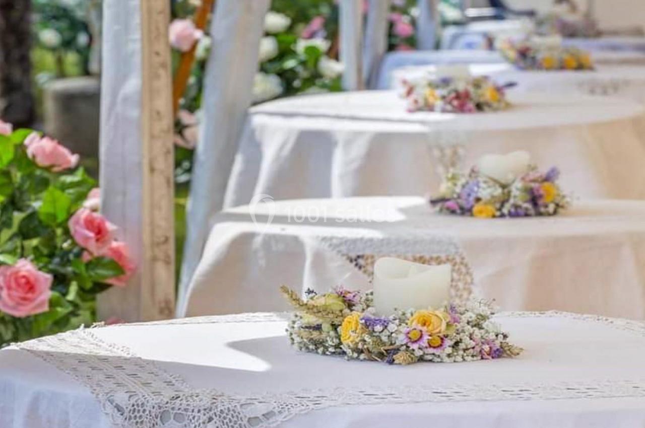 Tables décorées de nappes blanches, ornées de bougies entourées de fleurs, dans un cadre extérieur fleuri.