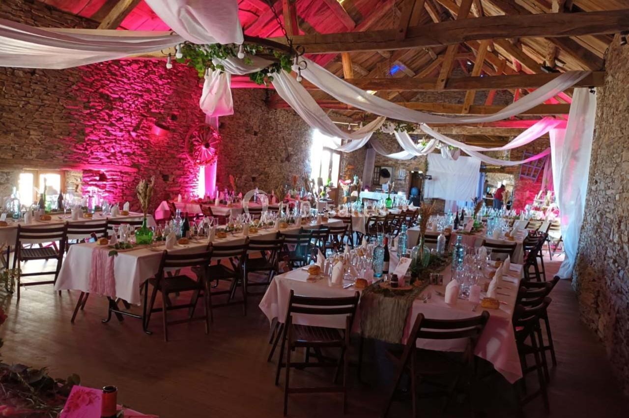 Salle de réception rustique décorée pour un événement, avec tables dressées, nappes blanches et éclairage rose.