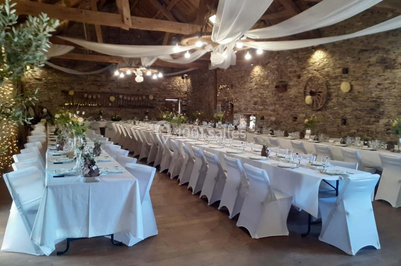Salle de réception décorée avec des tables alignées, nappes blanches et guirlandes lumineuses sous un plafond en bois.