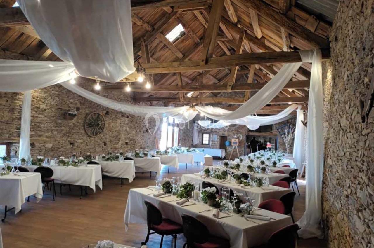 Salle de réception rustique avec tables décorées de nappes blanches, fleurs et chaises, sous un toit en bois avec voilages.