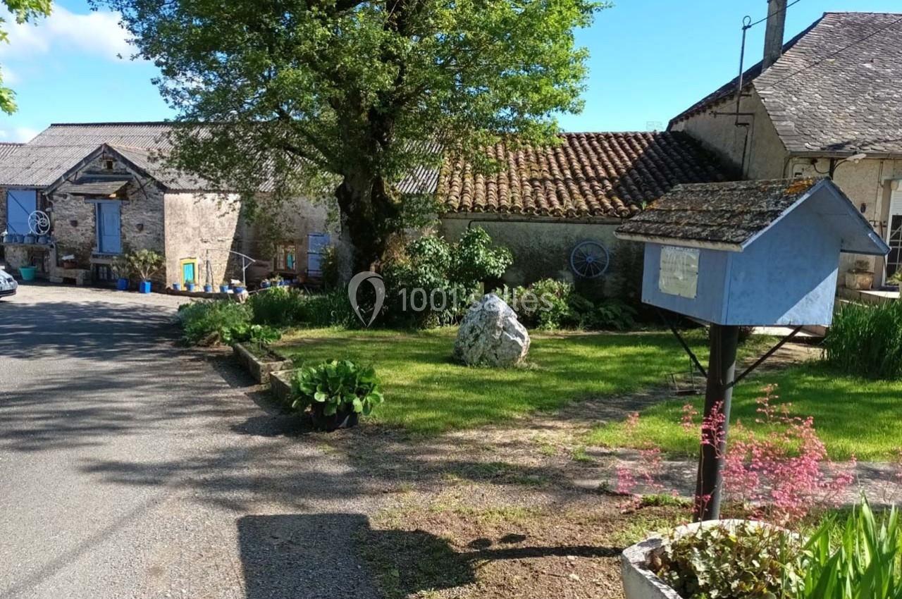 Cour de ferme avec bâtiments en pierre, boîte aux lettres bleue au premier plan et arbre au centre.