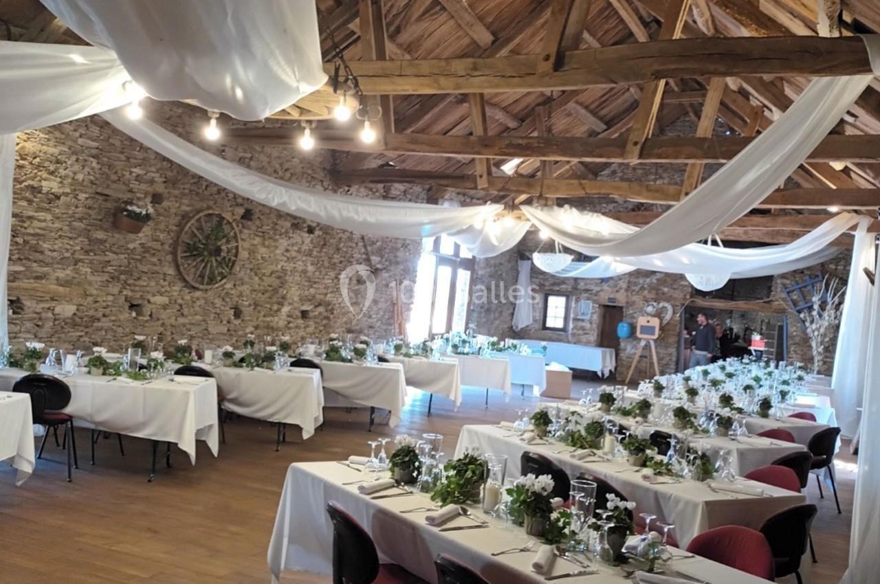 Salle de réception rustique avec tables dressées, nappes blanches et décorations végétales sous une charpente en bois.