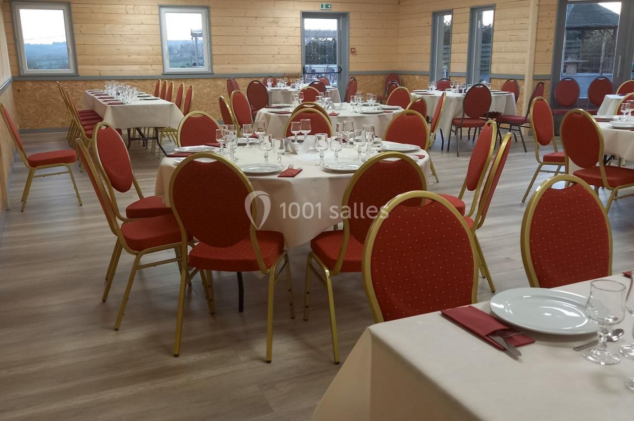 Salle de réception avec tables rondes dressées, nappes blanches, serviettes rouges et chaises rouges sur un sol en bois.
