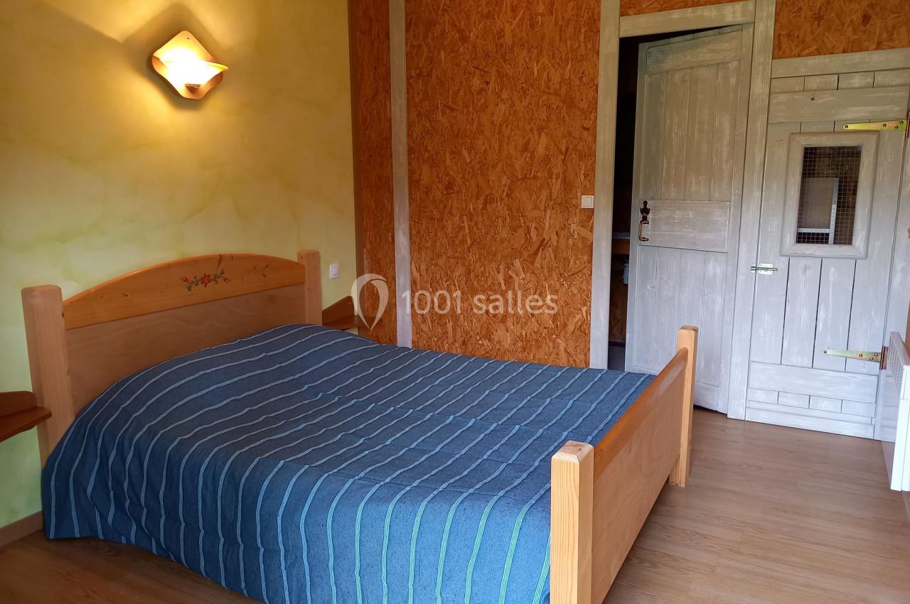 Chambre avec un lit double en bois, couverture bleue rayée, murs en bois et plâtre, éclairage mural allumé.