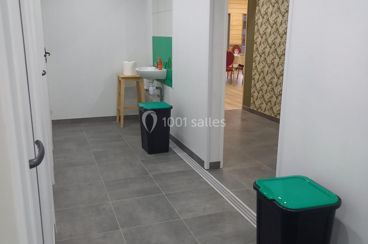 Couloir avec sol en carrelage gris, deux poubelles à couvercle vert, un lavabo et un mur décoré en arrière-plan.
