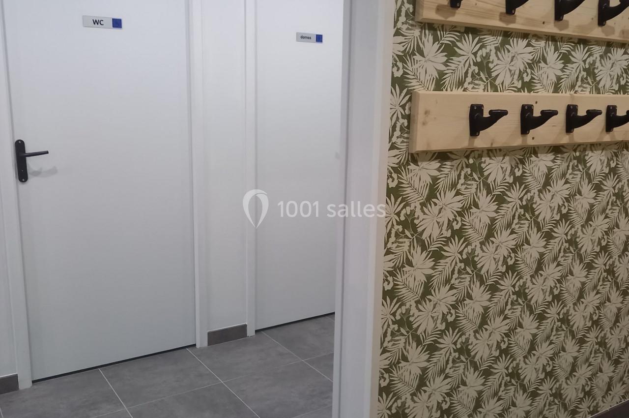 Vue d'un couloir avec des portes blanches de toilettes et un mur décoré de papier peint à motifs végétaux, équipé de patères.