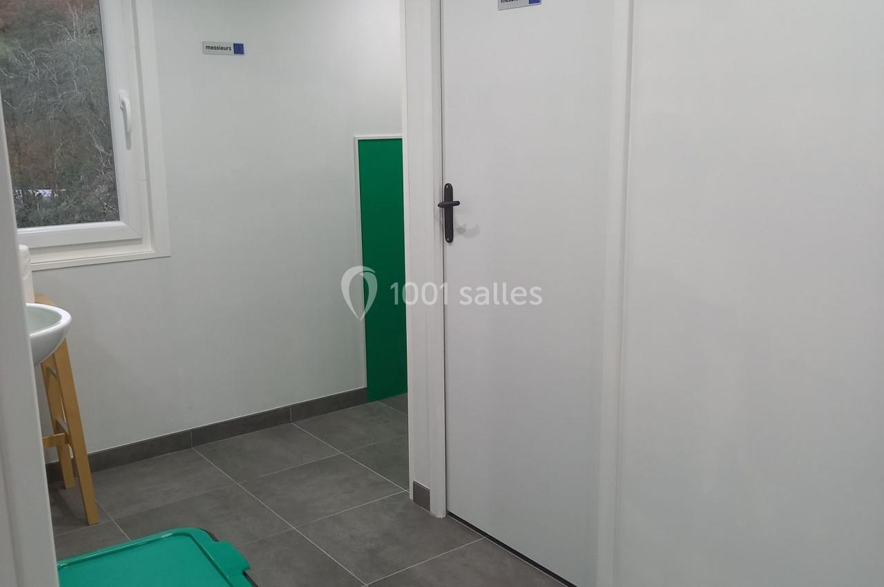 Vue d'un couloir avec sol en carrelage gris, une porte blanche fermée, une poubelle verte et une fenêtre.