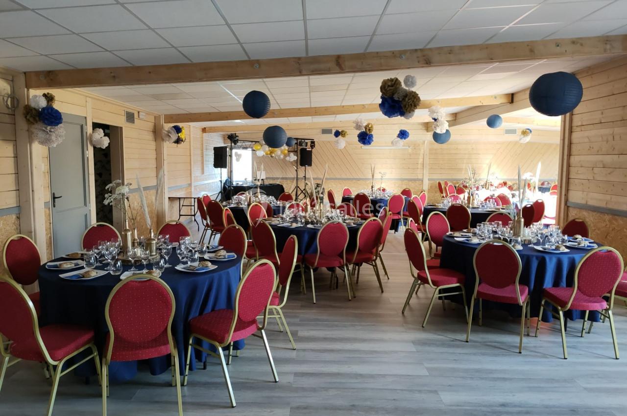 Salle décorée pour un événement, avec tables rondes dressées, nappes bleues, chaises rouges et décorations suspendues.