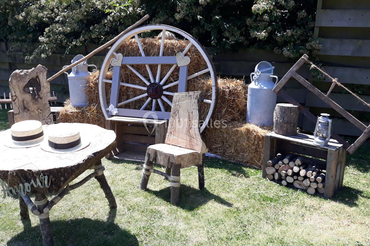 Décor champêtre avec table, chaises en bois brut, roue de charrette, bottes de paille et objets rustiques.