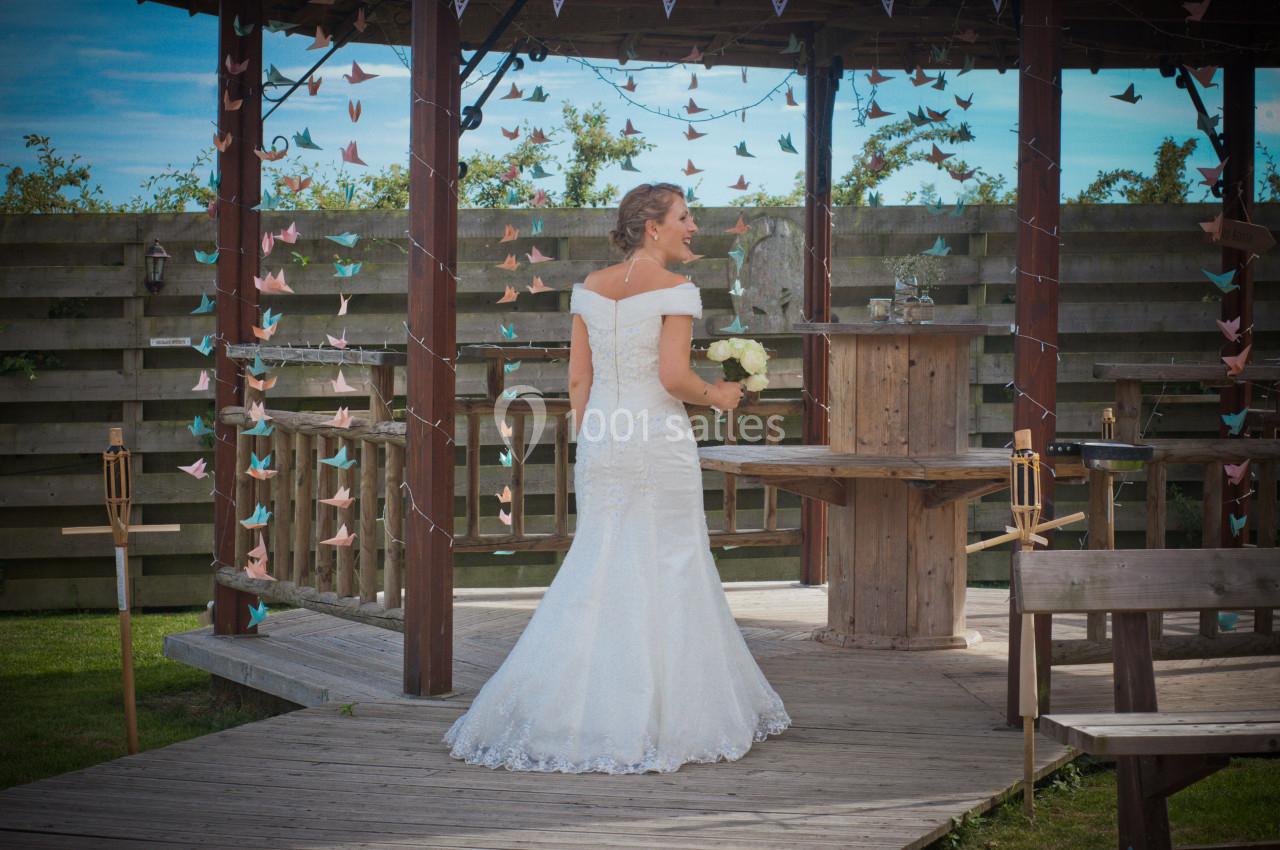 Une femme en robe de mariée blanche tient un bouquet de fleurs, debout sur une terrasse en bois décorée.