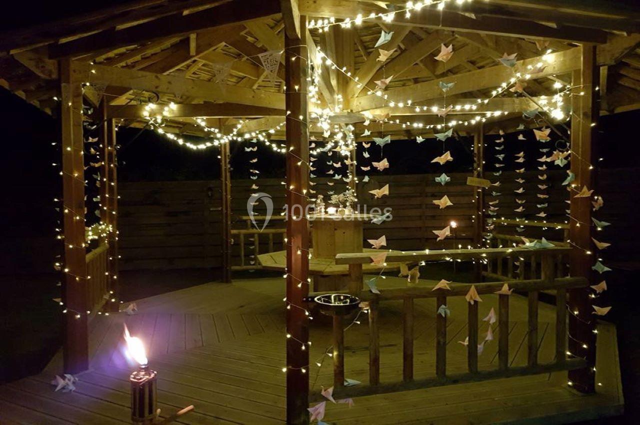 Kiosque en bois décoré de guirlandes lumineuses et d'origamis, éclairé par une torche dans une ambiance nocturne.