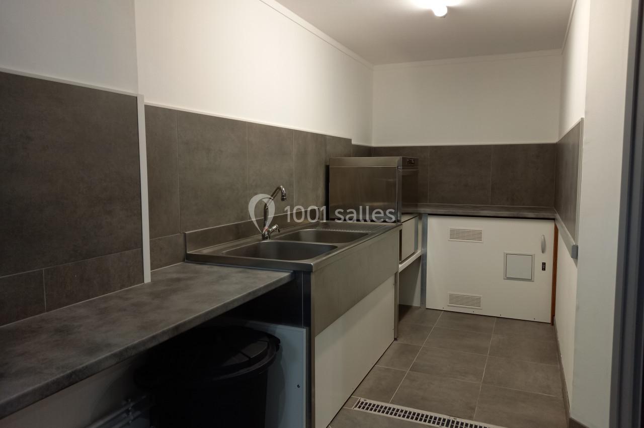 Cuisine professionnelle avec évier inox, plan de travail gris, carrelage mural et sol gris, éclairée par une lampe au…