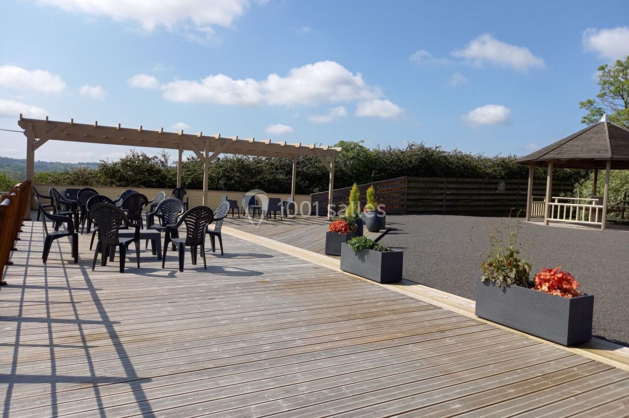 Terrasse en bois avec pergola, chaises noires, jardinières fleuries et kiosque en arrière-plan sous un ciel dégagé.